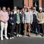 A group of sixteen people, both men and women, stand together outside in front of a building and CERTUS Pest control vehicles in Las Vegas. They are dressed casually and smiling, posing for a group photo in the bright sunlight.