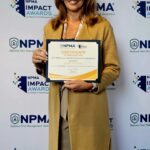 Dawn Hammond stands in front of an NPMA Impact Awards backdrop, smiling and holding an impactful award certificate with both hands. She is wearing a tan blazer and matching pants.