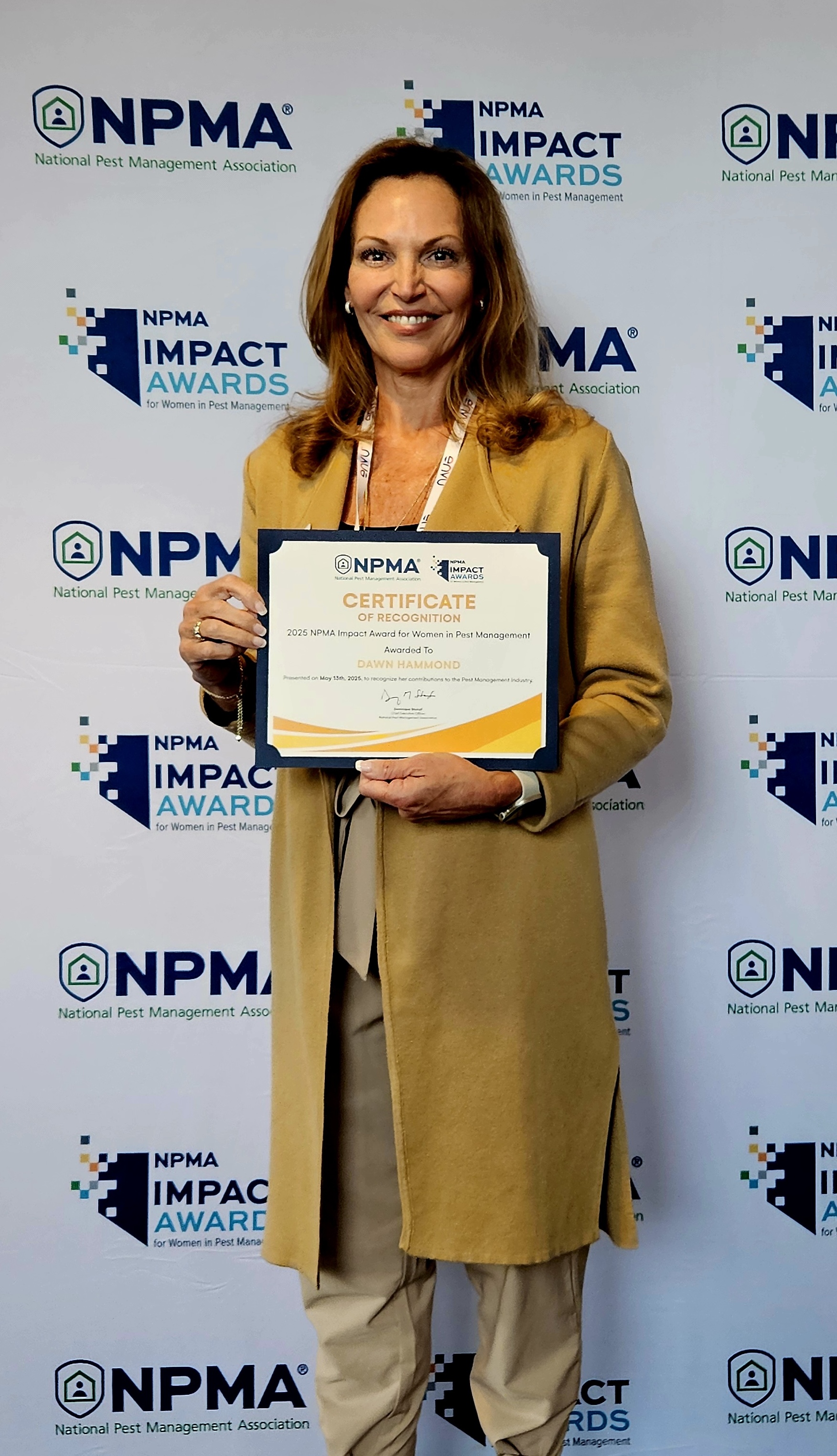 Dawn Hammond stands in front of an NPMA Impact Awards backdrop, smiling and holding an impactful award certificate with both hands. She is wearing a tan blazer and matching pants.